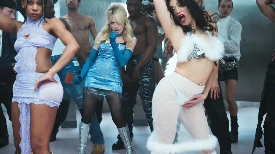 Madonna in a blue vinyl mini dress and long gloves dancing with performers in a music video scene.