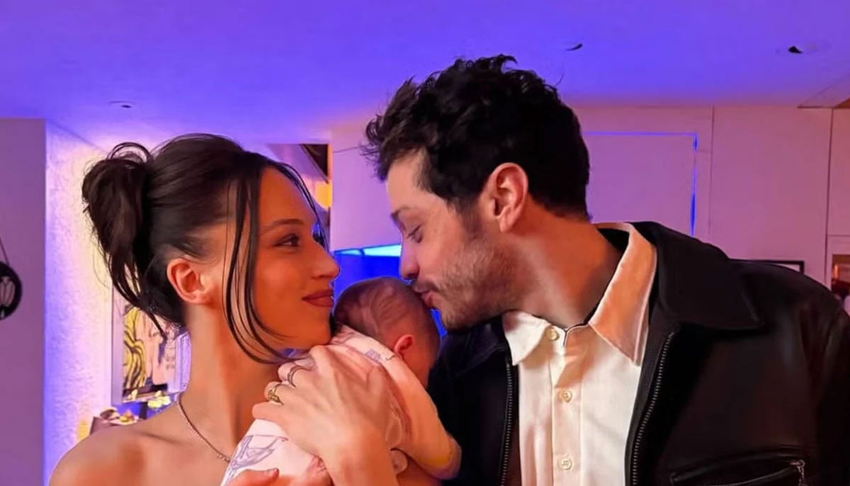Pete Davidson and Elsie Hewitt their newborn baby indoors under purple and blue ambient lighting, both looking at the infant.