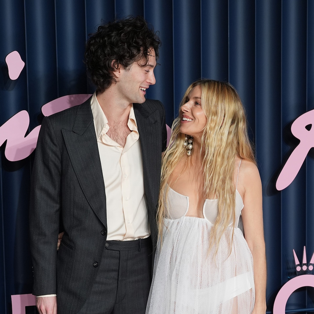 Oli Green and Sienna Miller Smiling at each other at a formal event with a blue curtain and pink decorative elements in the background.