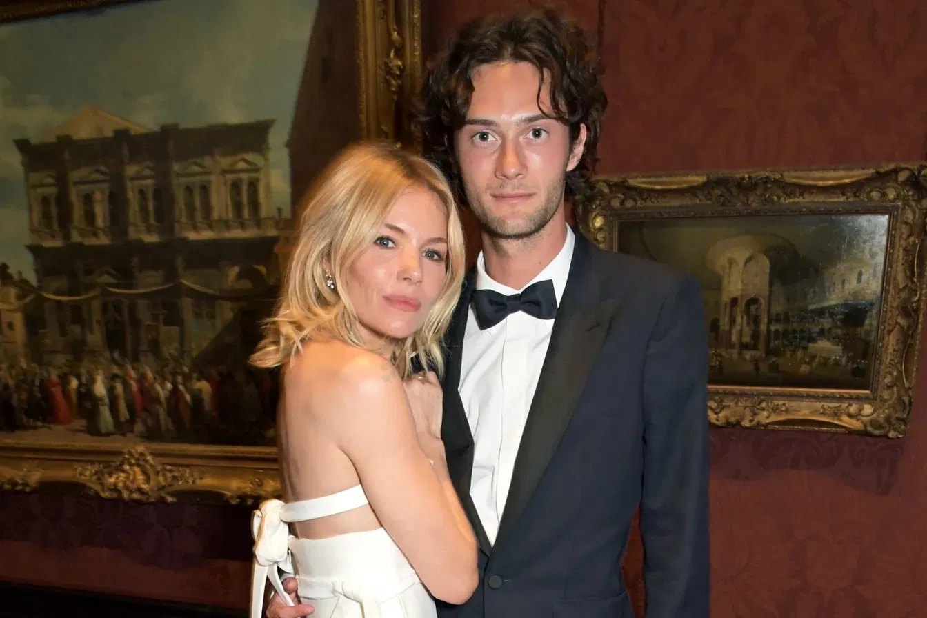 Sienna Miller and Oli Green posing together at a formal event with oil paintings on the wall behind them.