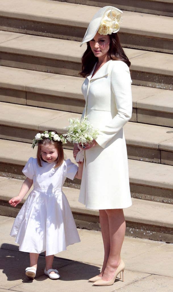Kate Middleton in a white coat dress and fascinator hat stands with a young girl in a white flower girl dress on stone steps.