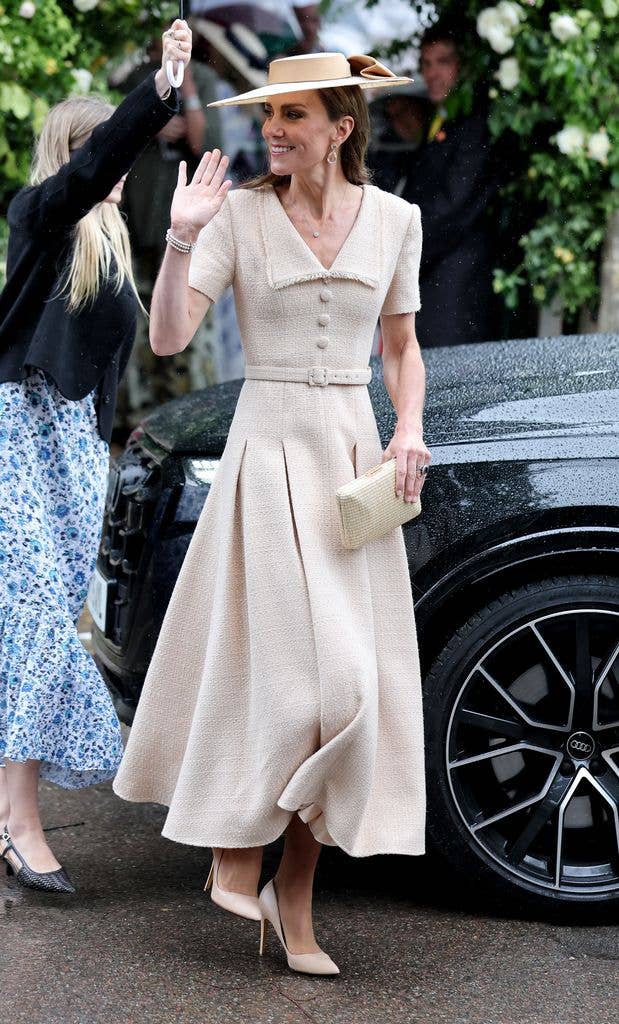 Kate Middleton in a cream-colored dress and wide-brimmed hat waves while standing beside a black car at an outdoor event.