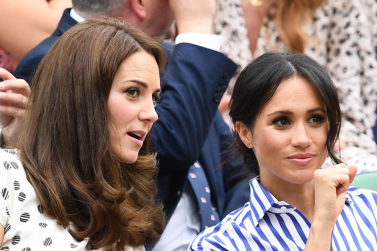 Kate Middleton and Meghan Markle sit together at what appears to be a formal event, one wearing a white patterned outfit and the other in a blue striped shirt.