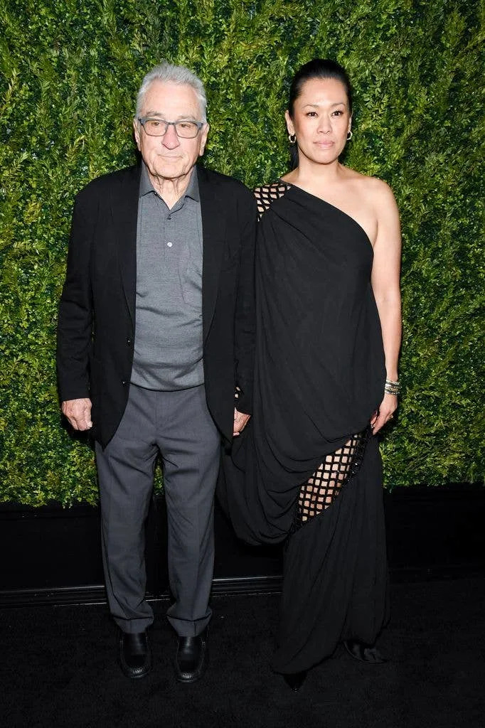 Robert De Niro and Tiffany Chen  posing together in front of a green foliage backdrop at a formal event.
