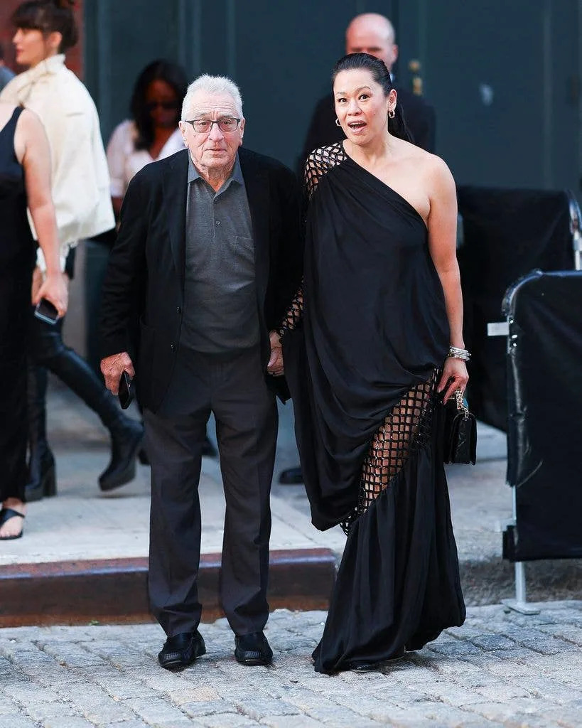 Robert De Niro in glasses and dark suit walks alongside Tiffany Chen in an elegant black draped gown at a formal event.