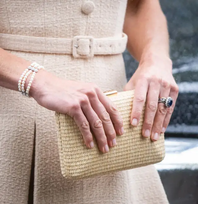 Close-up of Kate Middleton's hands holding a woven straw clutch, wearing a sapphire engagement ring and pearl bracelet with a beige belted dress.