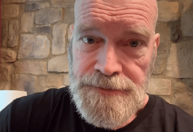 Tyler Mane with a full gray beard wearing a black shirt, photographed against a brick wall background.