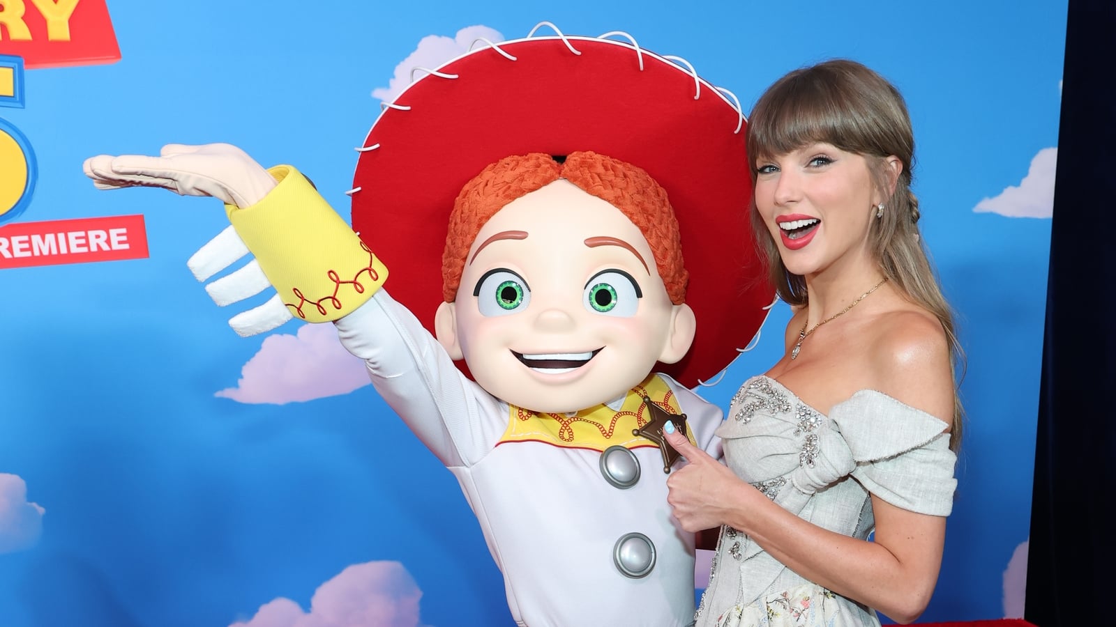 Taylor Swift in a gray off-shoulder dress poses with Jessie the cowgirl character at a movie premiere event.