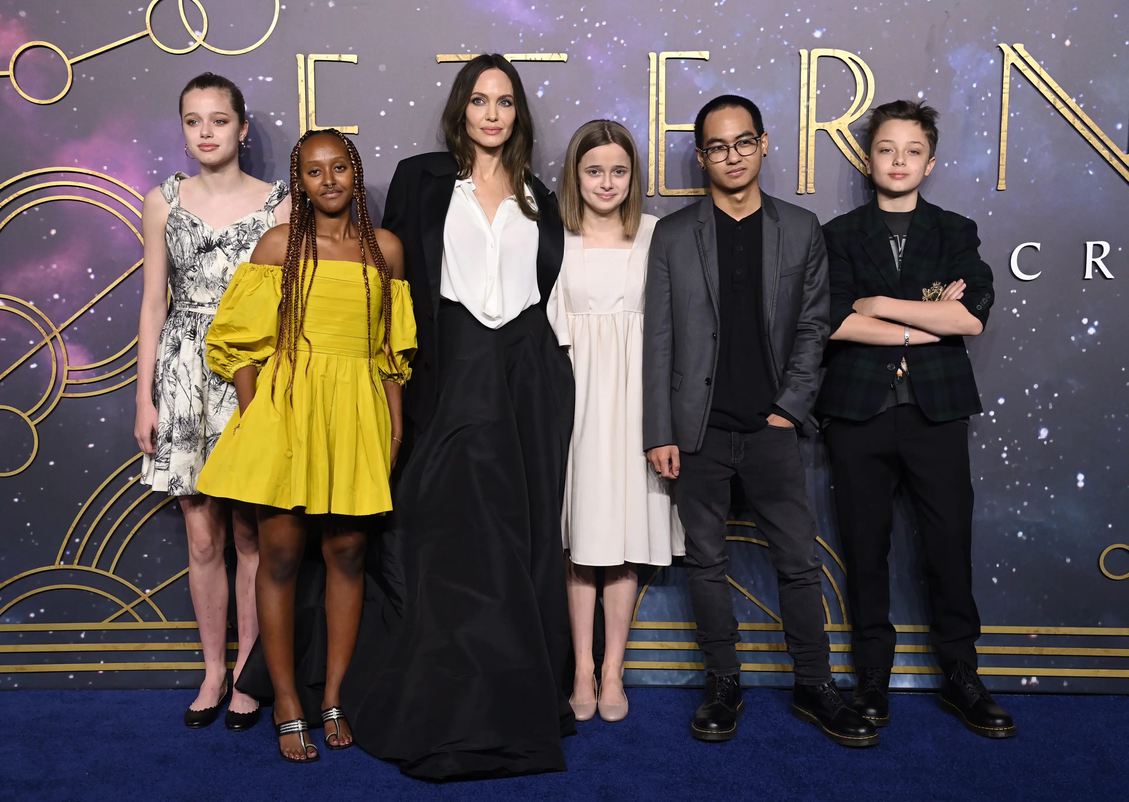 Angelina Jolie poses with her five children together at movie premiere for Eternals with a starry purple and gold backdrop behind them.