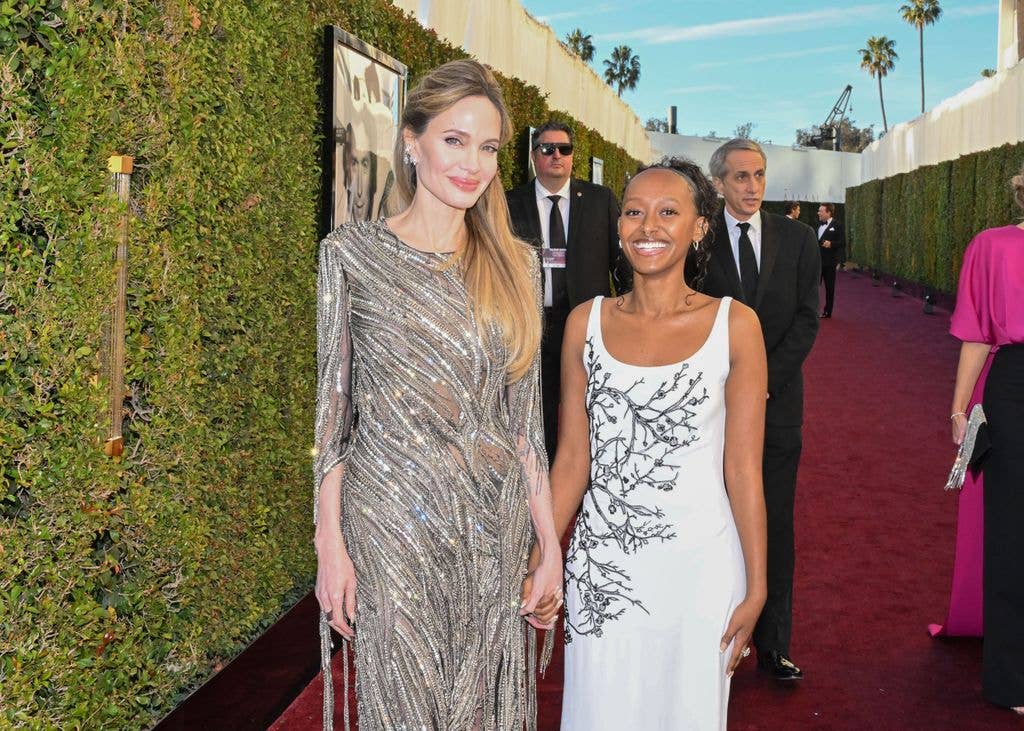 Angelina Jolie and Zahara Jolie holding hands on a red carpet at an event, with ivy-covered walls and security personnel in the background.