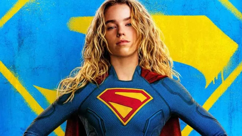 Milly Alcock with long wavy blonde hair wearing a blue Superman suit with red cape against a blue and yellow background.