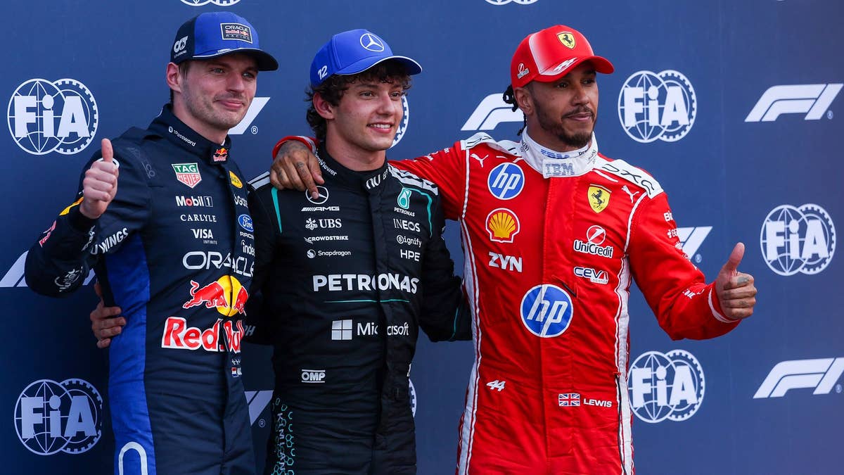 Max Verstappen, Kimi Antonelli and Lewis Hamilton on the podium wearing racing suits giving thumbs up gestures against an FIA backdrop.