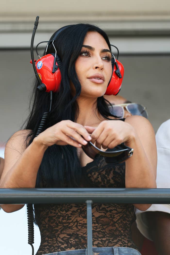 Kim Kardashian wearing red headphones with microphone, holding gaming controller, looking away thoughtfully.