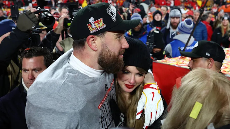 Travis Kelce in gray shirt stands with Taylor Swift in a black beanie at a crowded stadium celebration.