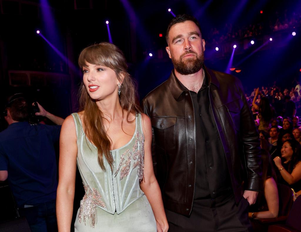 Taylor Swift and Travis Kelce stand together at an indoor event with purple lighting and a crowd visible in the background.