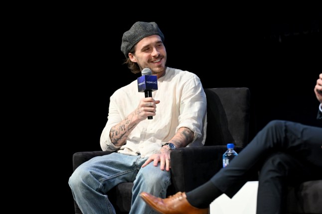 Brooklyn Beckham wearing a gray beanie and white shirt holds a microphone while seated on stage during what appears to be a panel discussion or interview.