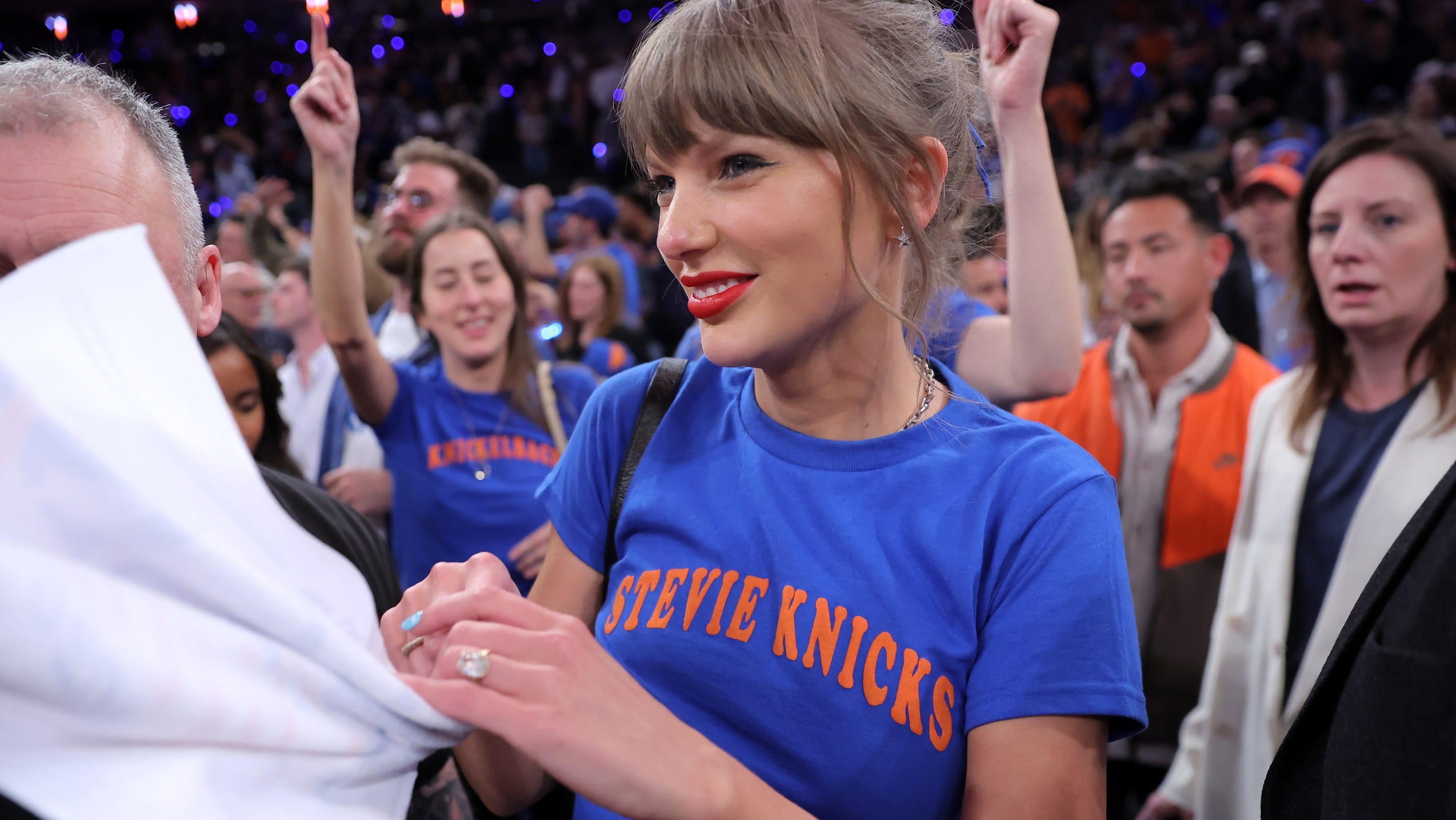 Taylo Swift in a blue Knicks shirt shakes hands with someone at a crowded sports arena event.