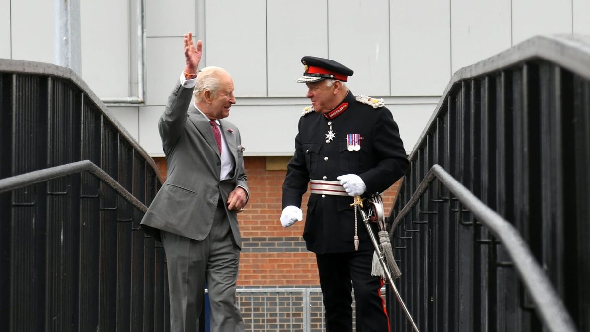King Charles in a gray suit raises his fist while speaking with a military officer in formal dress uniform on a metal bridge.
