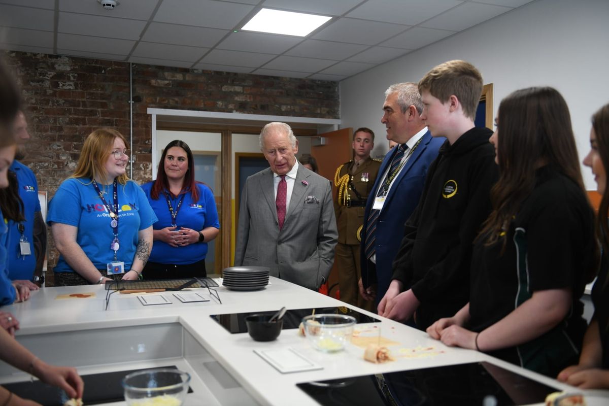 King Charles in a gray suit speaks with young staff members in blue uniforms during a kitchen facility tour.