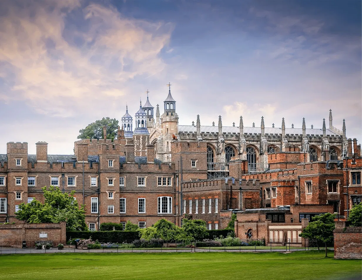 Eton College's historic red brick buildings and Gothic chapel towers against a cloudy sky, viewed from the playing fields.