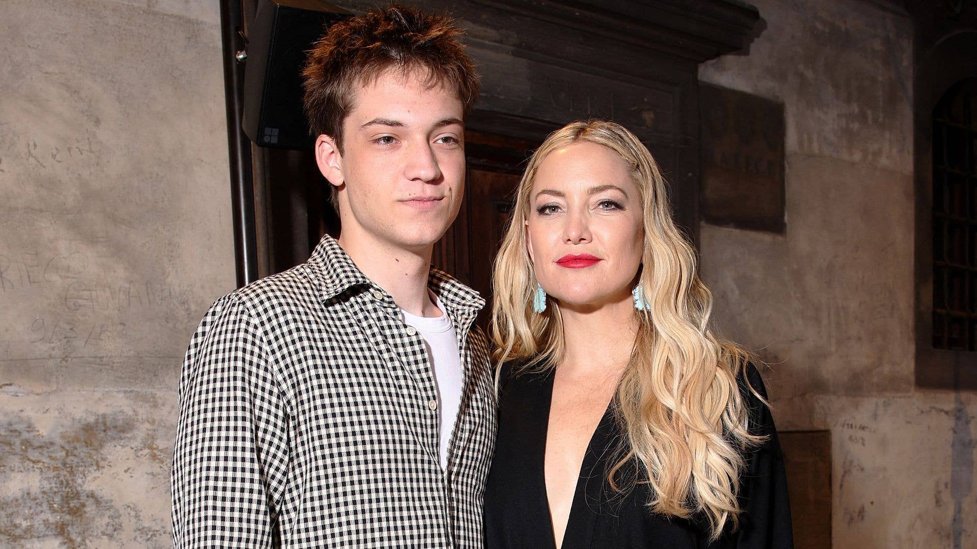 Ryder Robinson and Kate Hudson standing together indoors against a concrete wall backdrop, one wearing a black and white checkered shirt, the other in black.