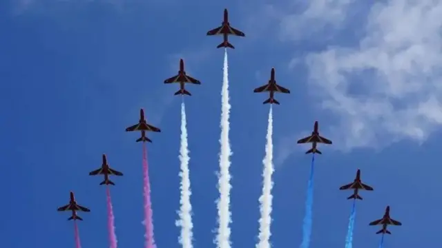Red Arrows Royal Air Force jets performing an aerobatic display with colored smoke trails in formation against a blue sky.