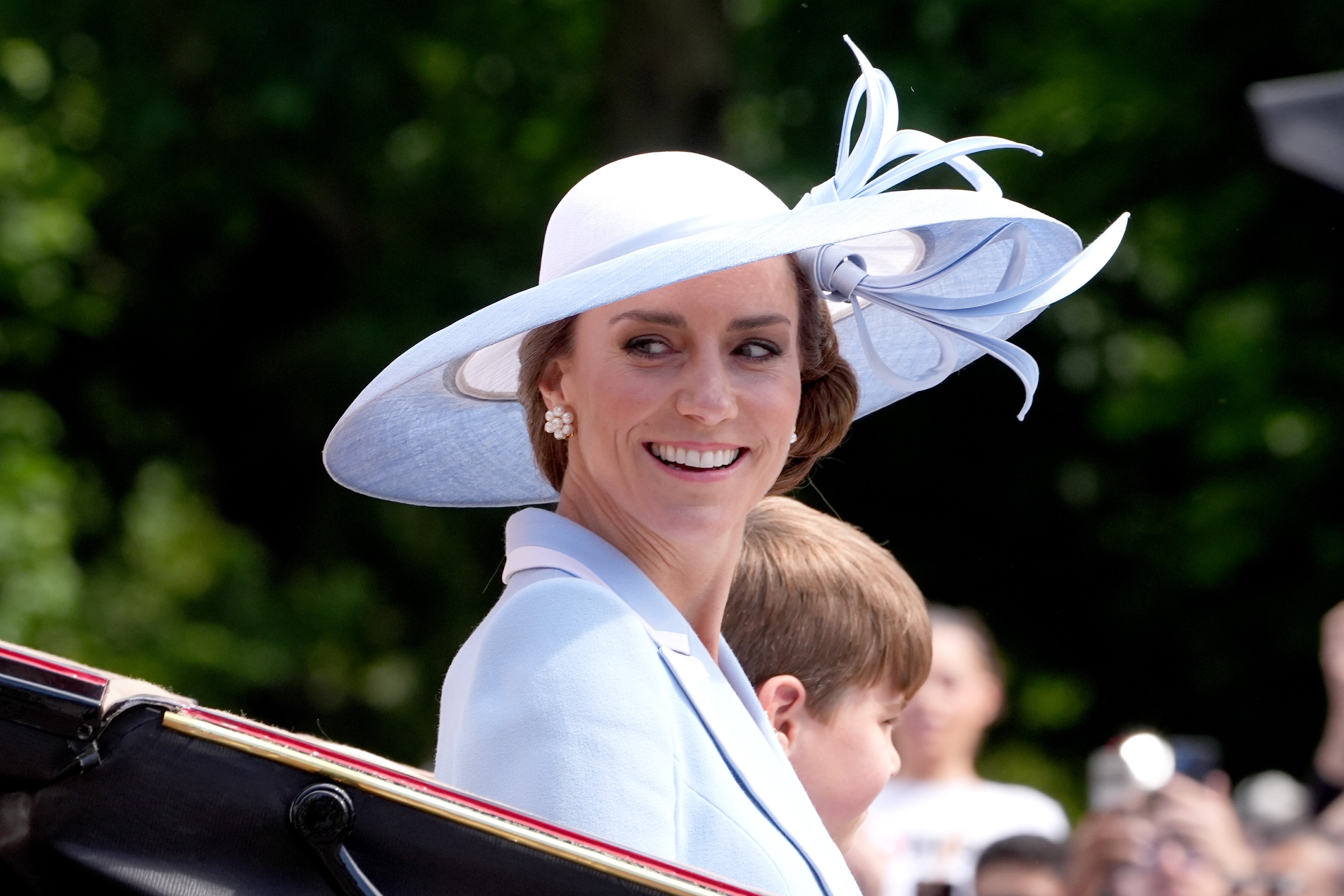 Kate Middleton in a light blue outfit and wide-brimmed hat with feather decoration smiles while riding in an open carriage.