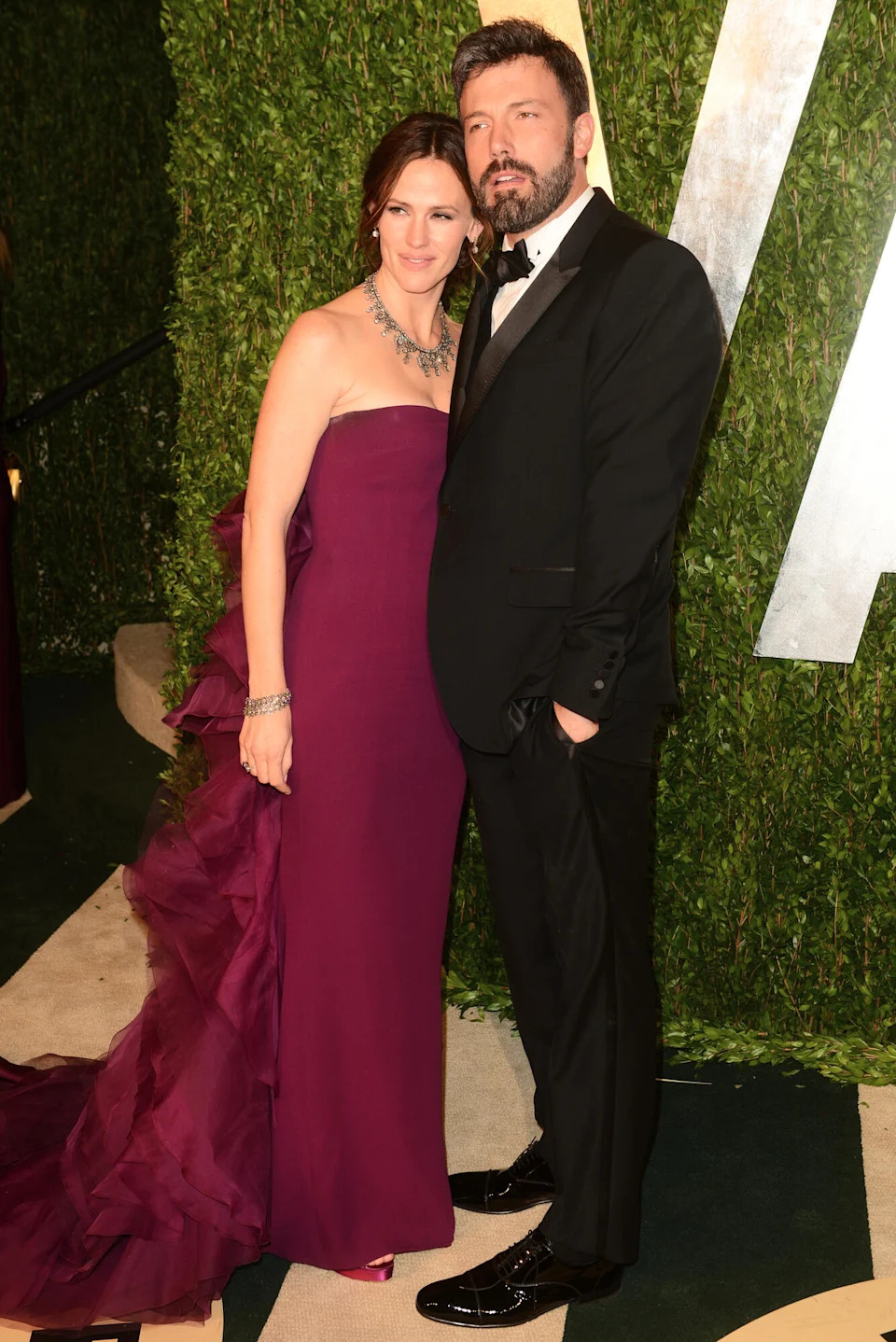 A couple in formal attire poses together at an event with ivy-covered walls in the background.