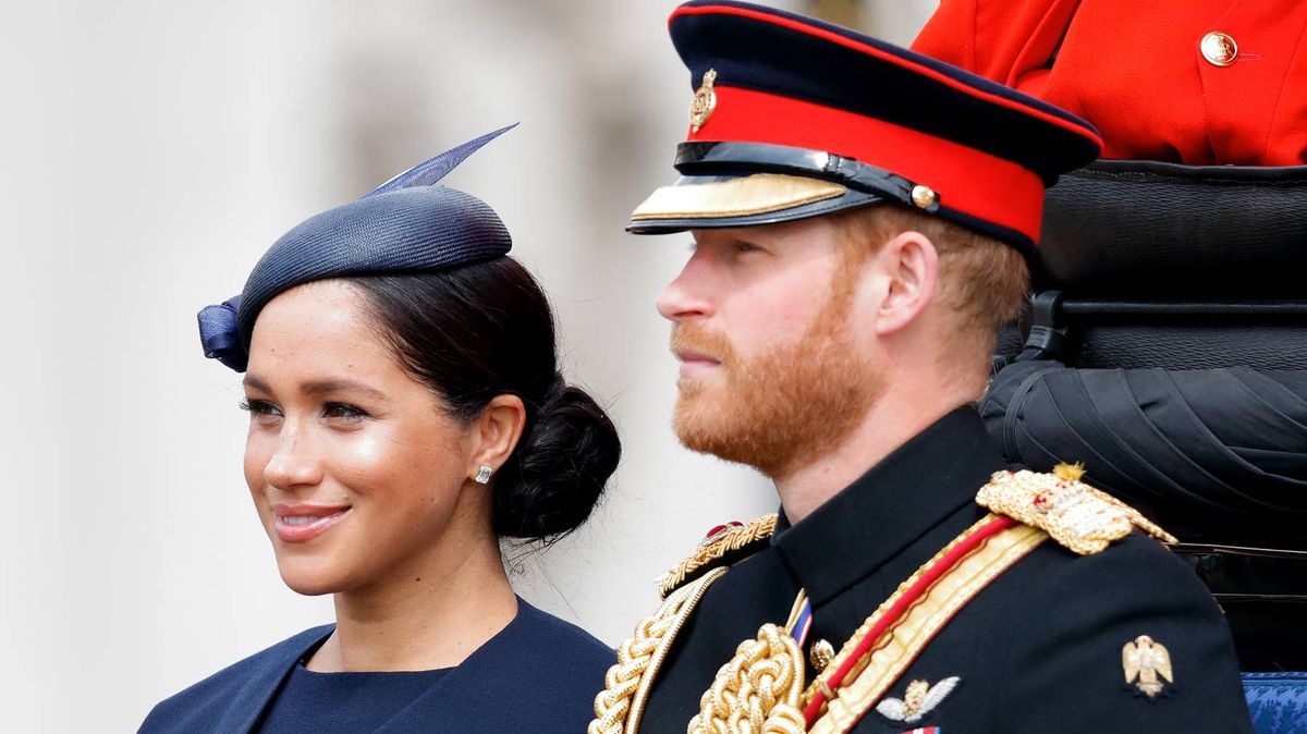 Meghan Markle in navy dress with fascinator hat stands beside Prince Harry in military dress uniform with red and black ceremonial jacket.