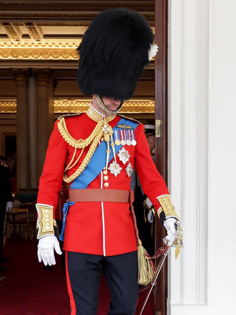 Prince William in full ceremonial dress uniform with bearskin hat, red tunic, blue sash, medals, and white gloves.