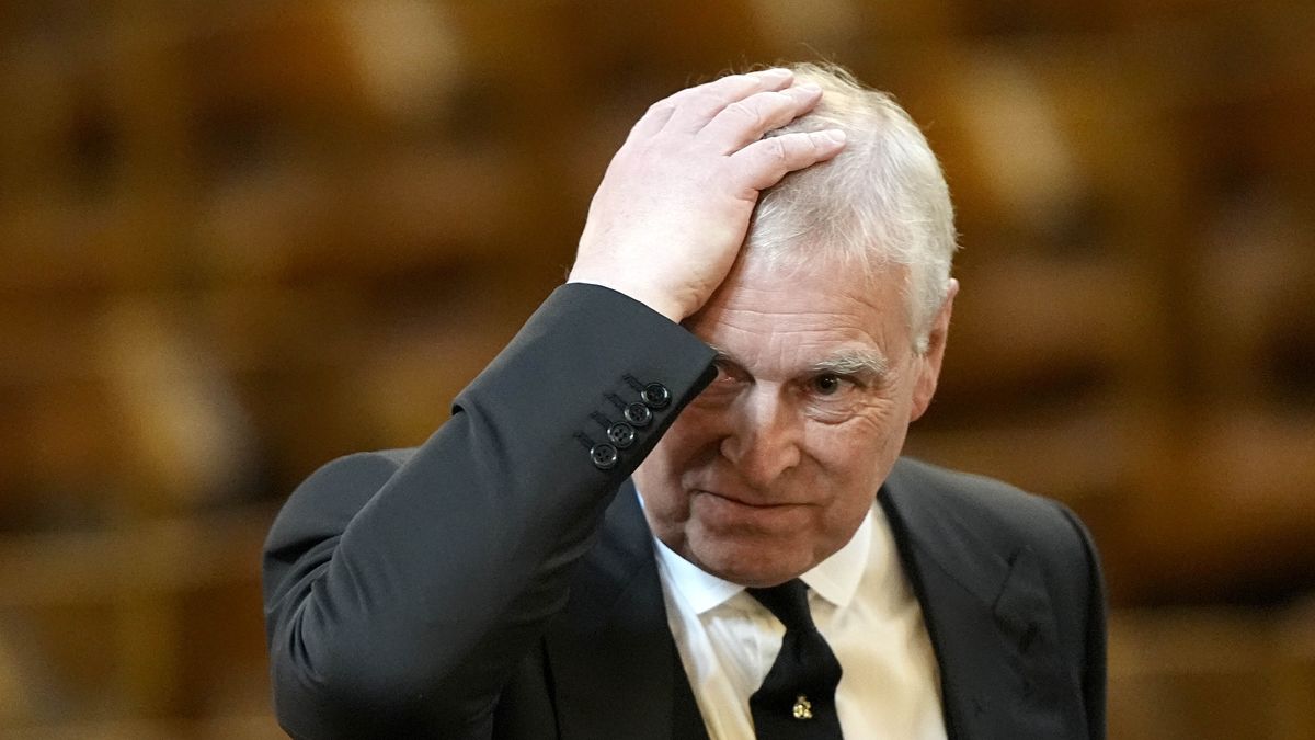 Prince Andrew in a dark suit and tie touches his head while smiling slightly at a formal event.