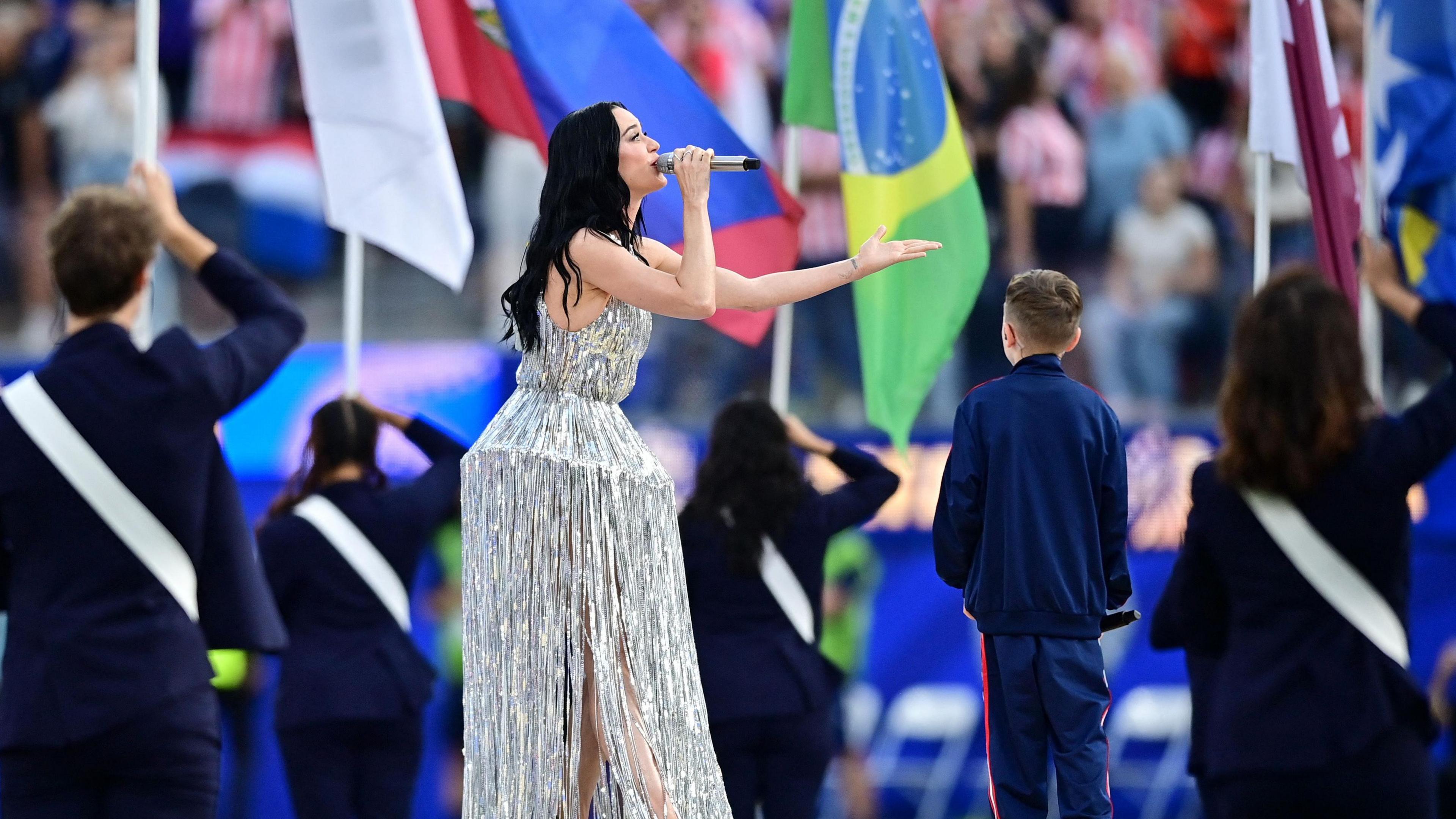 Katy Perry in a silver fringe gown sings into a microphone surrounded by international flags and uniformed attendants.
