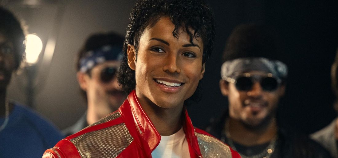 Jaafar Jackson as Michael Jackson in a red and gold jacket smiles at the camera while others stand behind him in a still from the film Michael