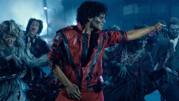 Jaafar Jackson as Michael Jackson in Michael wearing a red leather jacket dances amid zombie-like figures in a dark for thriller music video shooting scene.