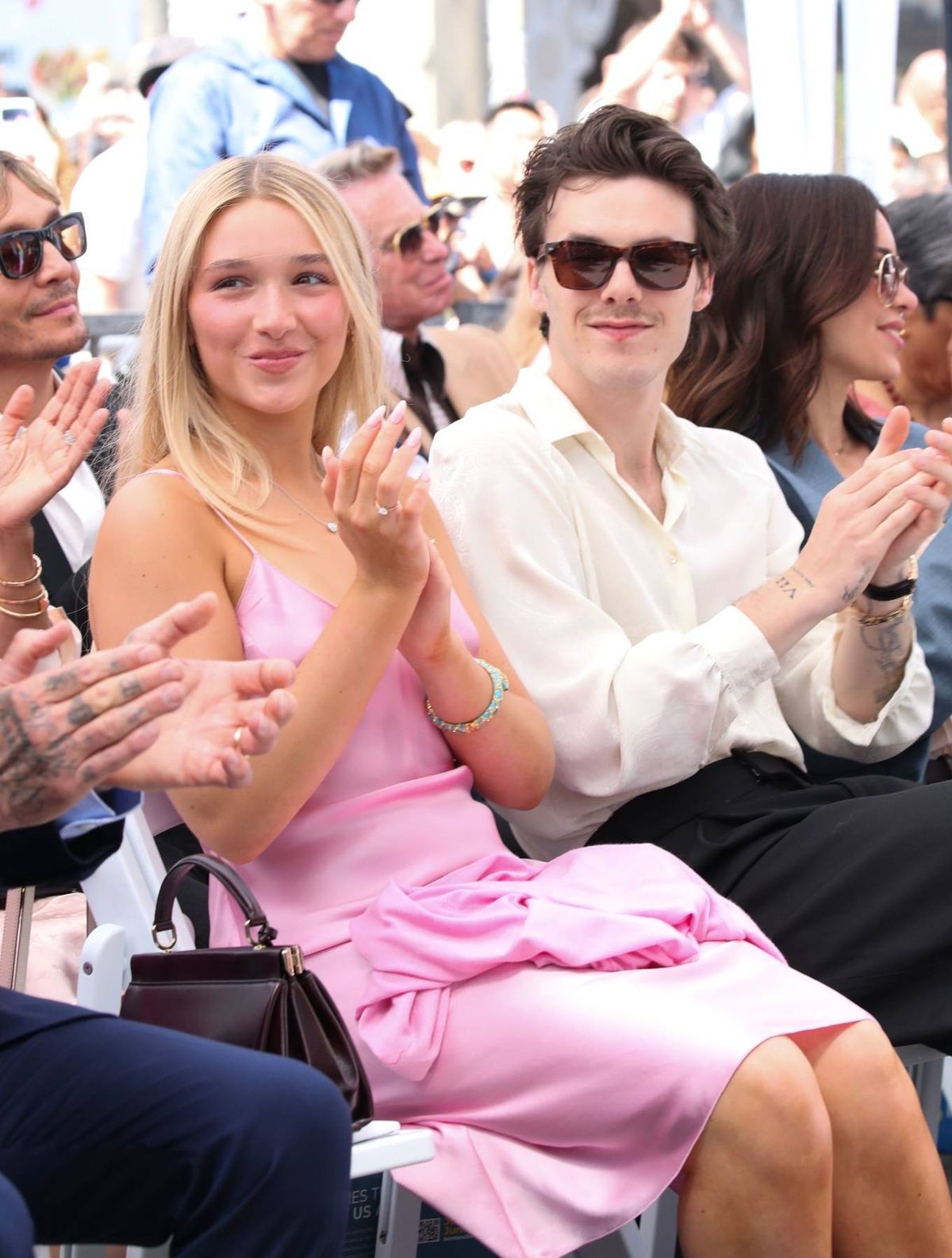 Harper Beckham in a pink dress and Cruz Beckham in a white shirt applaud at an outdoor event surrounded by other attendees.