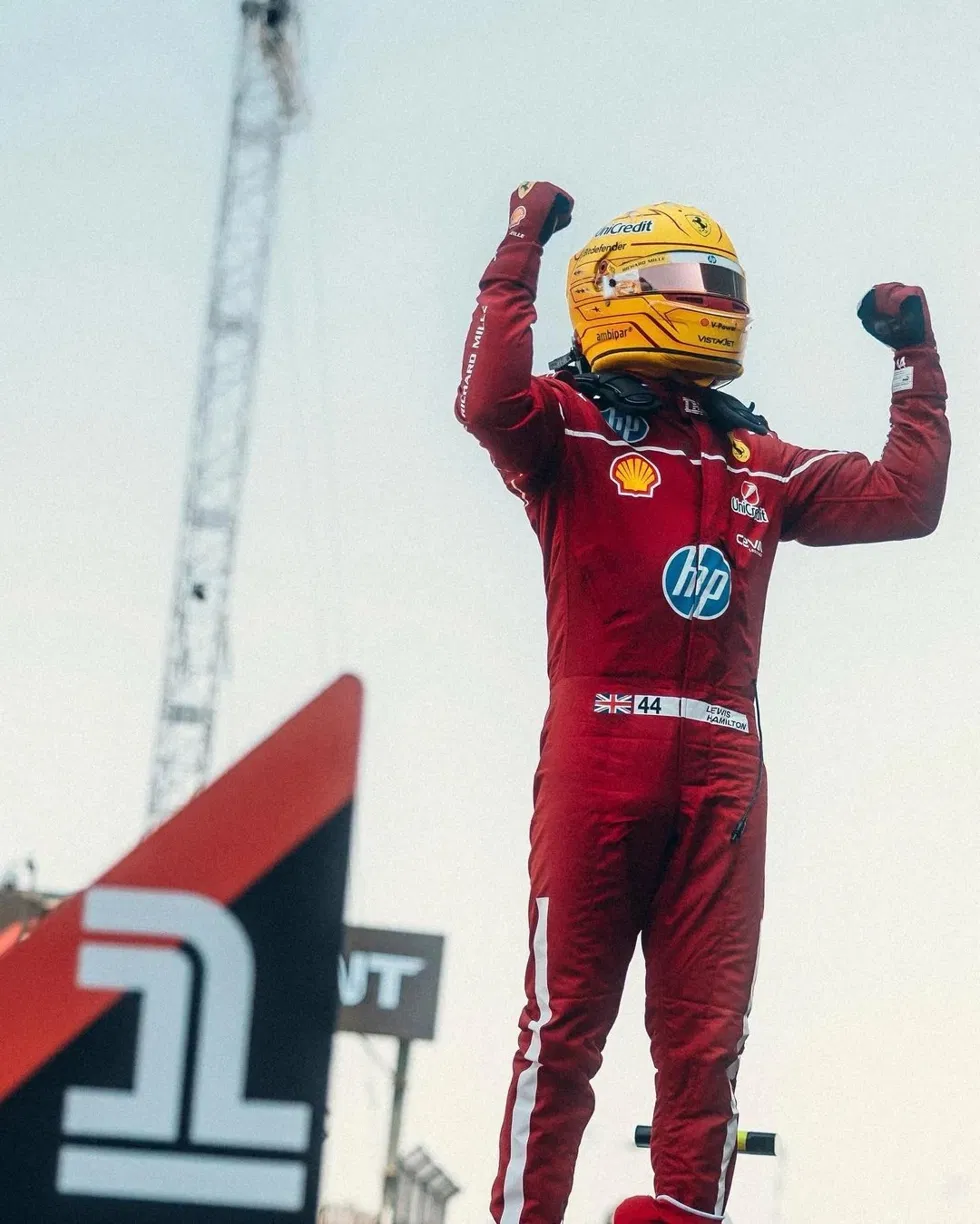 Lewis Hamilton in his Ferrari race suit and helmet, raising both arms in celebration.