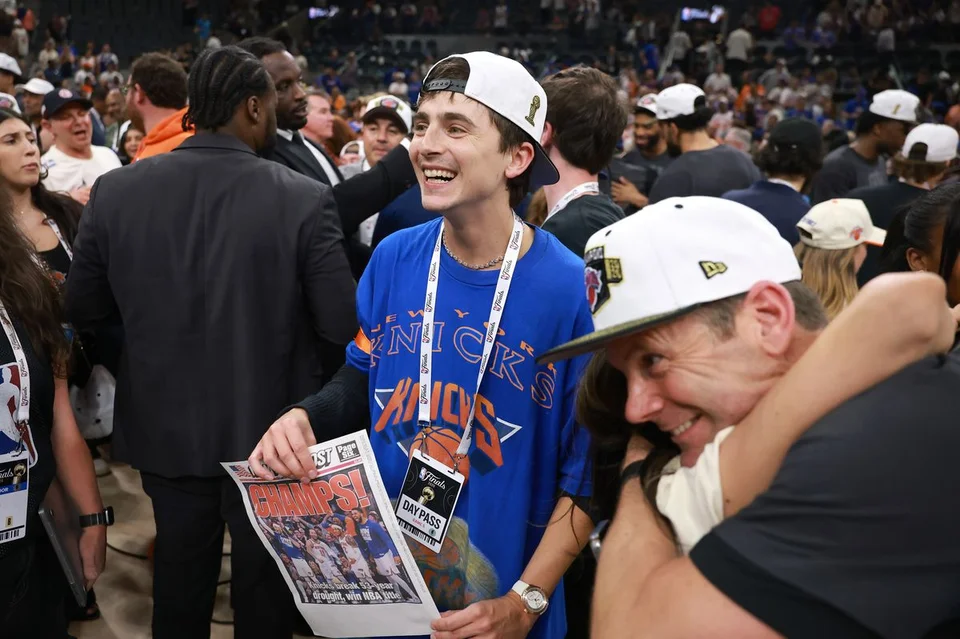 Timothée Chalamet wearing a New York Knicks jersey and cap, holding a newspaper with the headline 
