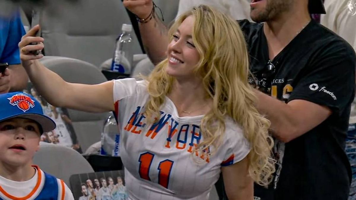 Sydney Sweeney in a New York Knicks jersey takes a selfie while surrounded by fans at what appears to be a basketball game.