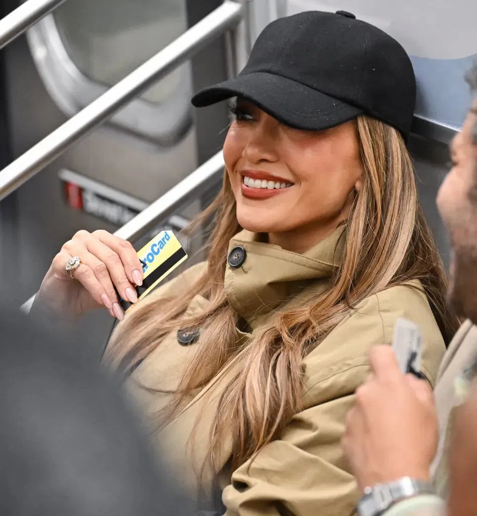 Jennifer Lopez wearing a black cap and tan jacket, smiling while holding a Metro card on a subway train.