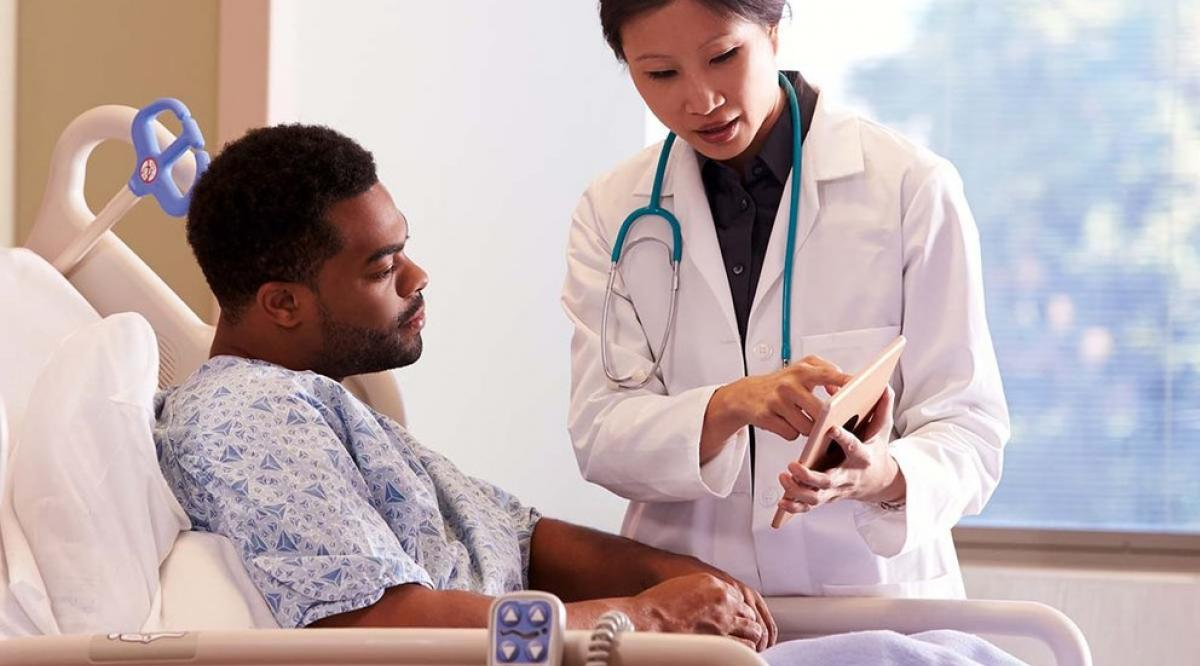 A doctor in a white coat showing a tablet to a patient lying in a hospital bed.