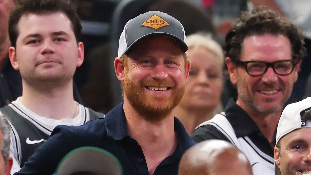 Prince Harry wearing a gray and white SBFD cap and dark polo shirt smiles while seated among other spectators at a sporting event