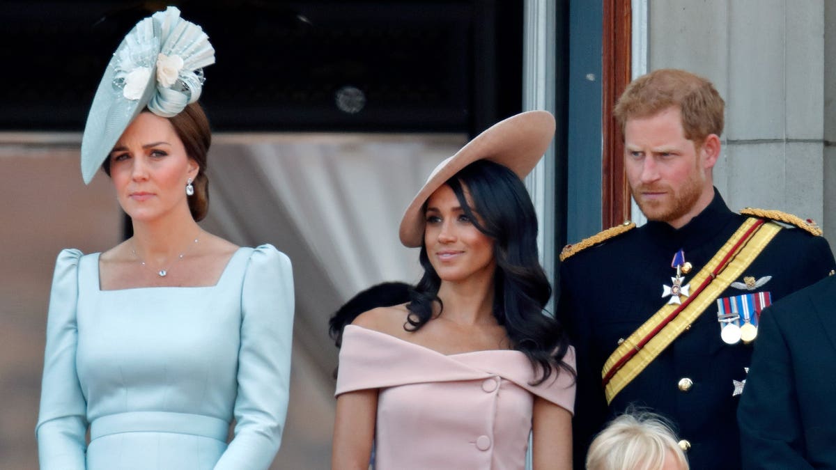 Kate Middleton, Meghan Markle and Prince Harry stand on a palace balcony: Kate in light blue with a fascinator, Meghan pink, and Harry in military dress uniform.