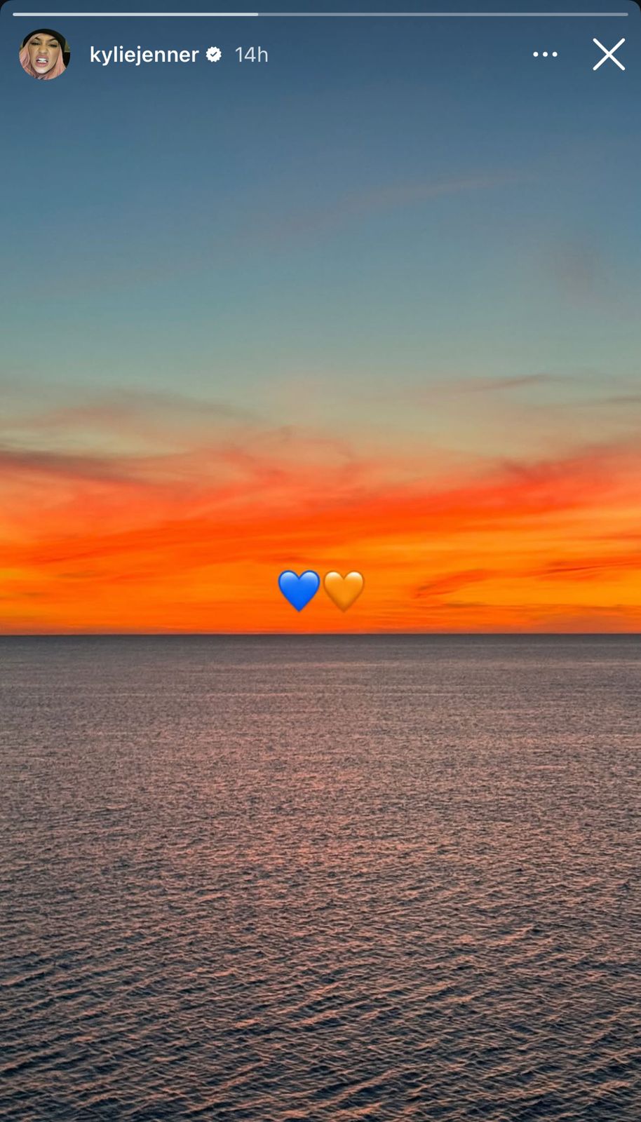 A screenshot of Kylie Jenner's Instagram story showing a sunset over the ocean with blue and orange heart emojis overlaid on the horizon.