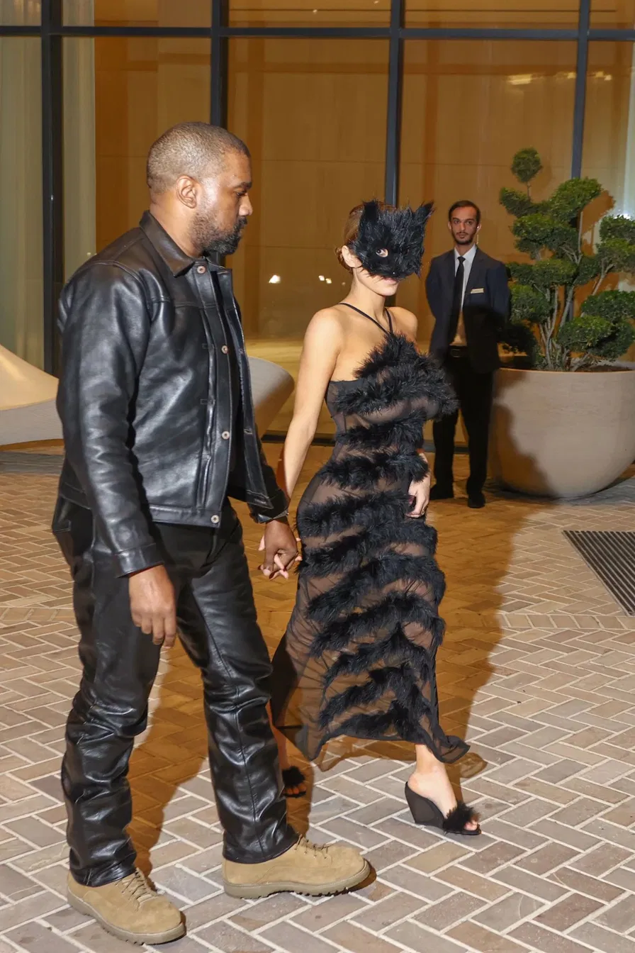 Bianca Censori in a cat mask and sheer dress holding hands with Kanye West wearing a black leather jacket at a public event
