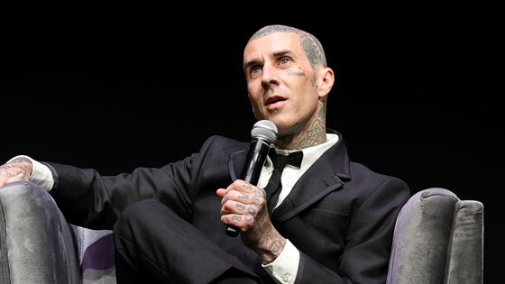 Travis Barker in a black tuxedo with a bow tie holds a microphone while seated, speaking at what appears to be a formal event.