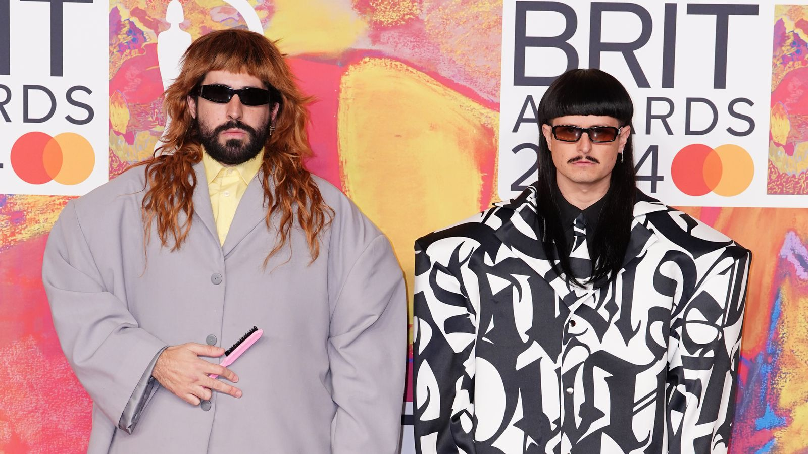 Robin Schulz with a long auburn wig and grey coat, and Oliver Tree with a black bob wig and graphic-print suit, at the Brit Awards 2024.