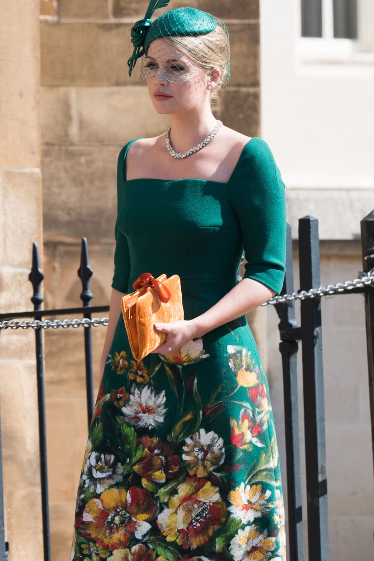 Lady Kitty Spencer wearing a green floral dress, matching fascinator hat with veil, and holding an orange clutch bag.