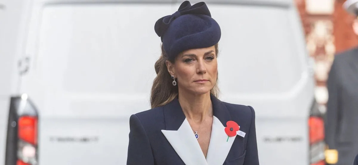 Kate Middleton in formal navy and white attire with a red poppy pin, wearing a navy fascinator hat with a bow.