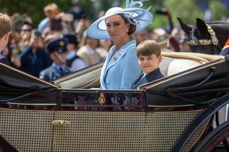 Kate Middleton in a light blue outfit and matching fascinator hat sits in an open carriage with Prince Louis in dark formal wear.