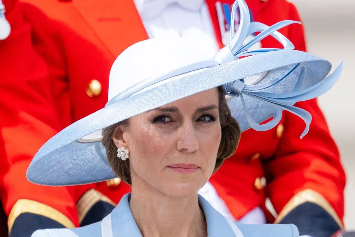 Kate Middleton wearing a pale blue wide-brimmed hat with ribbon detail and pearl earrings, with guardsmen in red uniforms in the background.